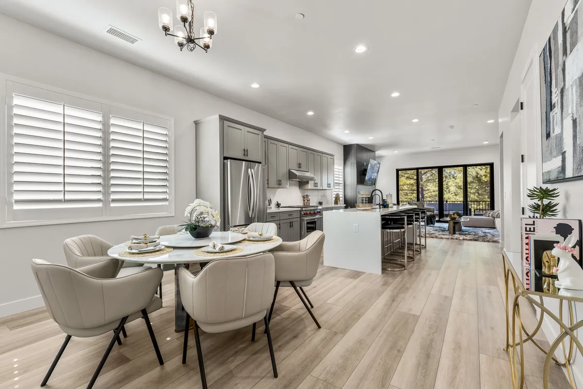 A stylish dining area is set with a large table, elegant tableware, and is adjacent to a kitchen island with modern appliances. Large windows bathe the space in natural light and offer glimpses of the outdoors, enhancing the open-plan feel.