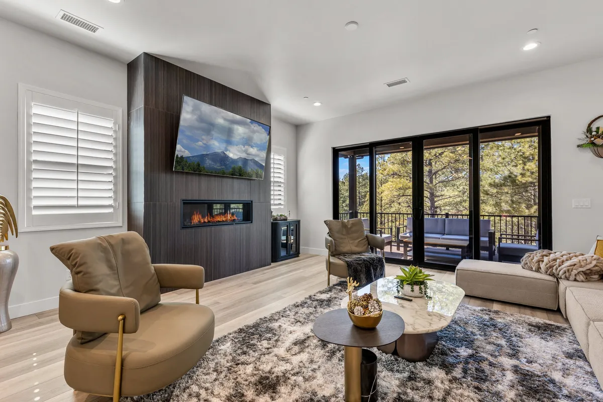 This image features a modern fireplace as the focal point, surrounded by comfortable seating and a large flat-screen TV. Large windows let in ample natural light and provide tranquil forest views, while a coffee table with decorative elements sits at the center, creating a welcoming and inclusive atmosphere.